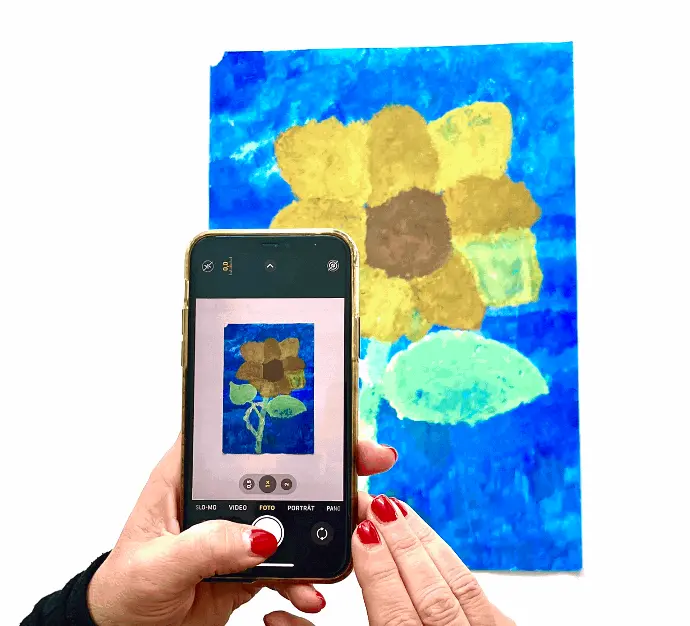 Frau fotografiert mit dem Smartphone ein Kinderkunstwerk, das eine gelbe Blume auf blauem Hintergrund zeigt.